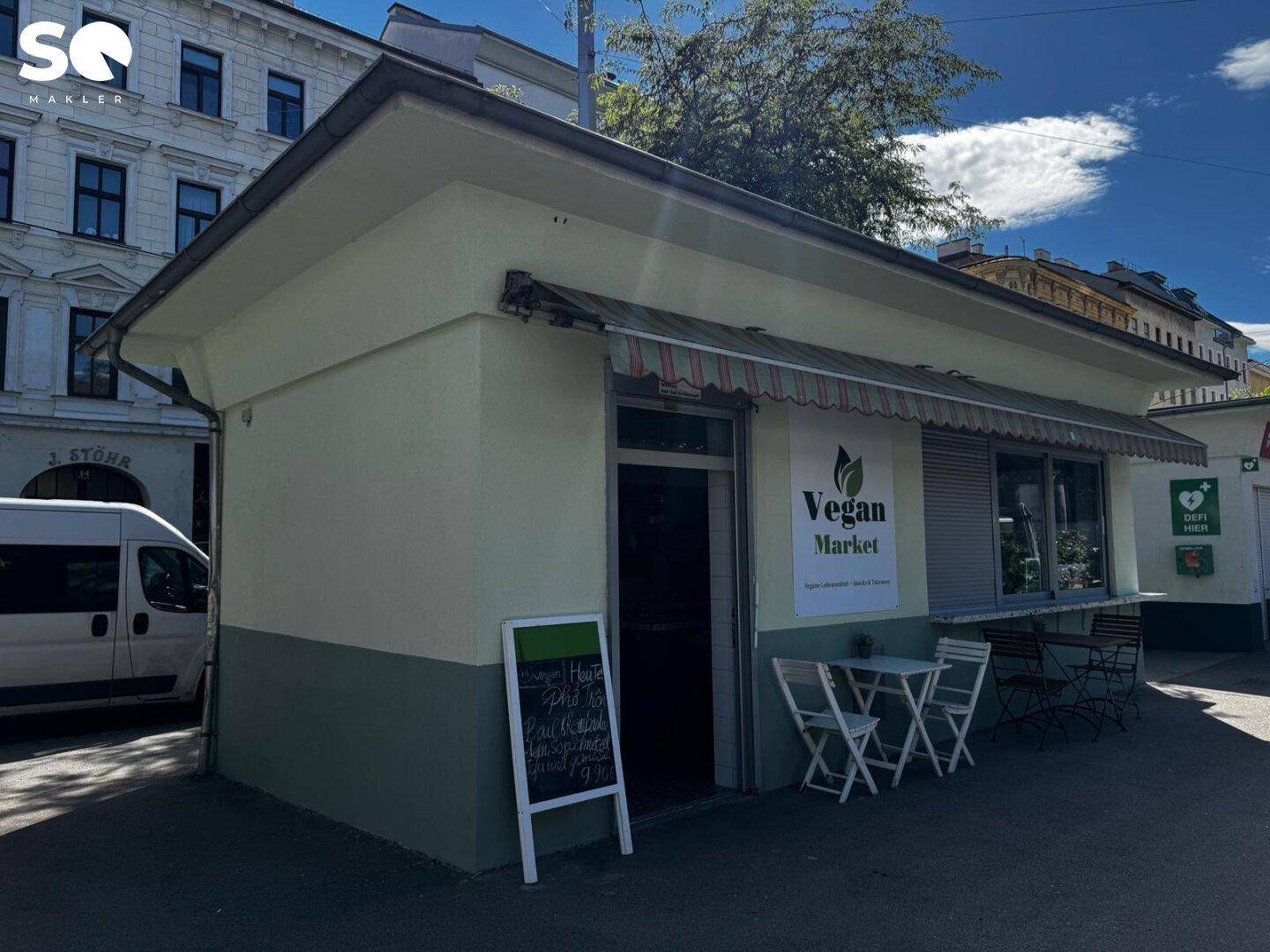 Ansicht des "Vegan Market" von der Seite, mit Außensitzplätzen und dem Eingangsbereich unter blauem Himmel.