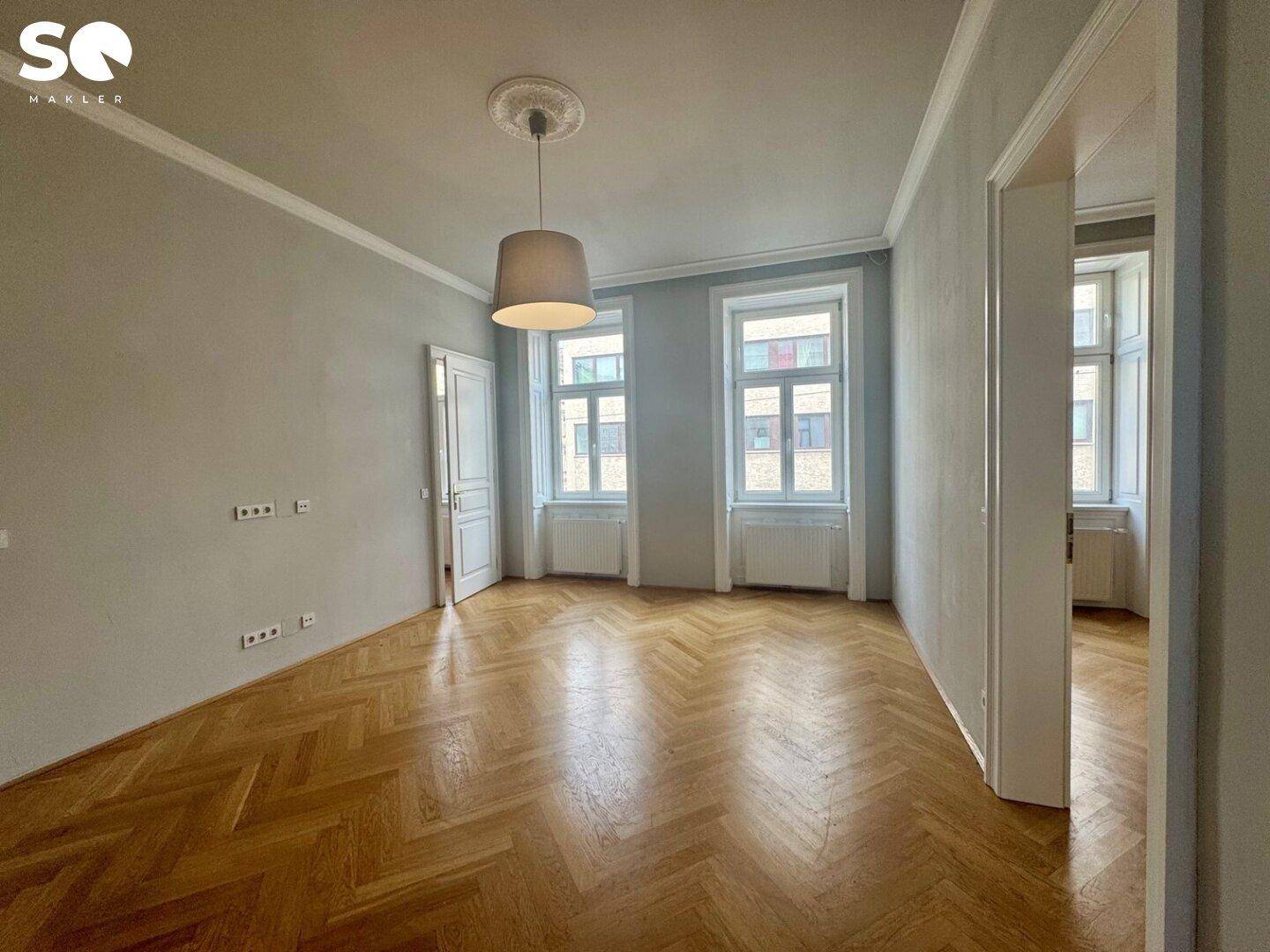 Geräumiges Zimmer mit elegantem Fischgrätparkett und zwei hohen Fenstern, die eine helle Atmosphäre schaffen.