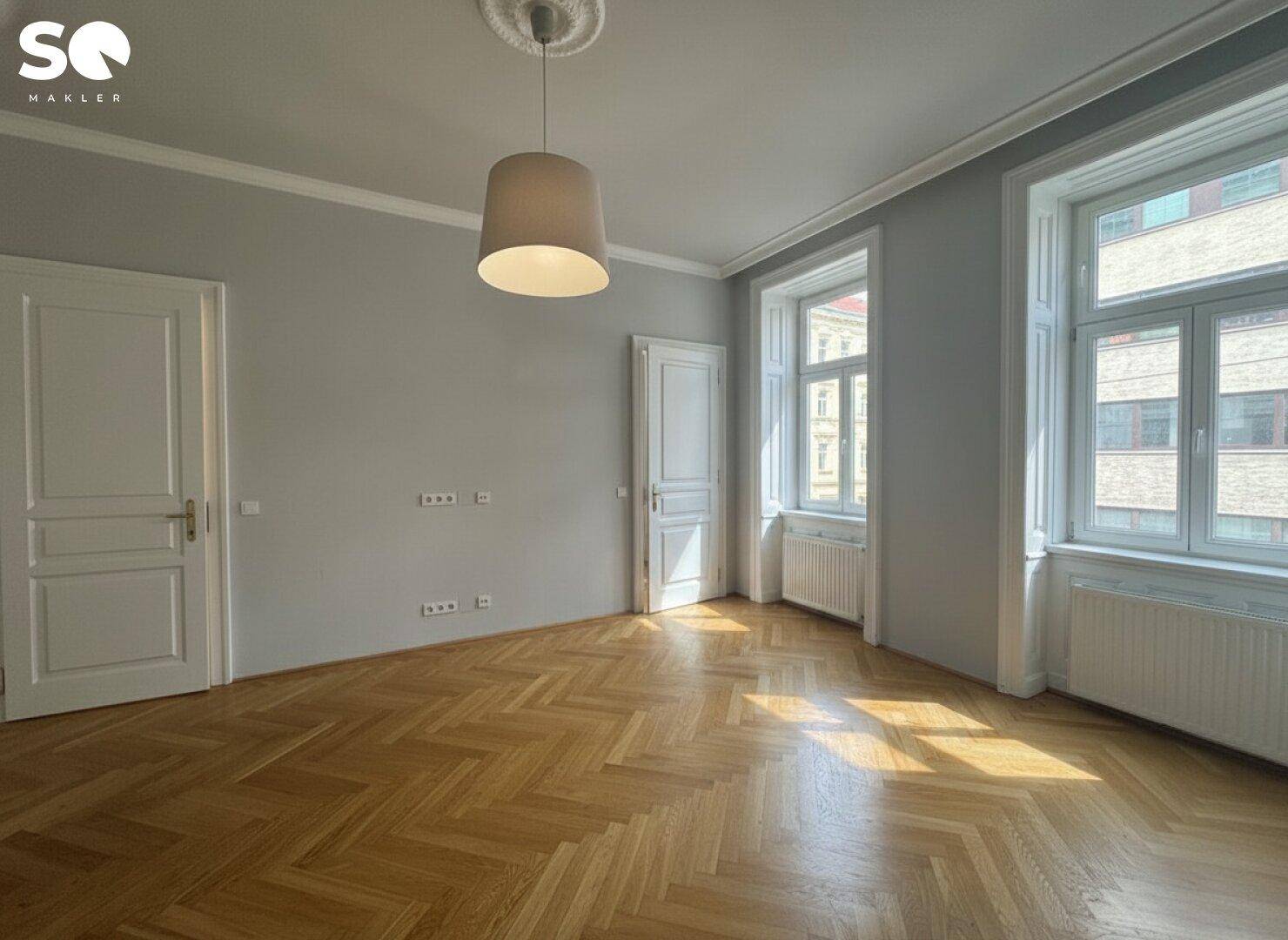 Lichtdurchflutetes Zimmer mit klassischem Fischgrätparkett und großen Fenstern, die viel Tageslicht hereinlassen.