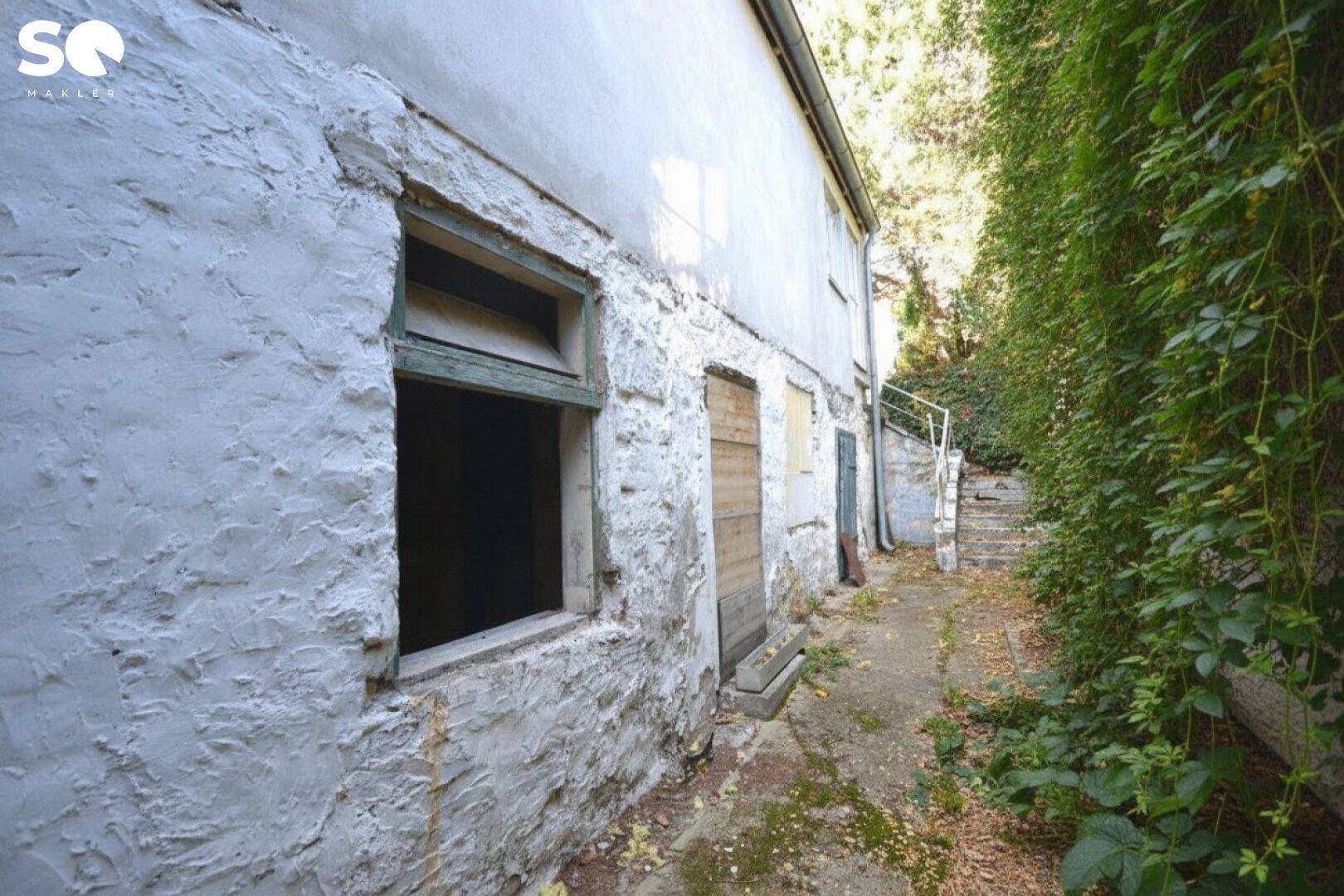 Altes Gebäude mit verputzter Fassade und zugewachsenem Weg im Außenbereich.