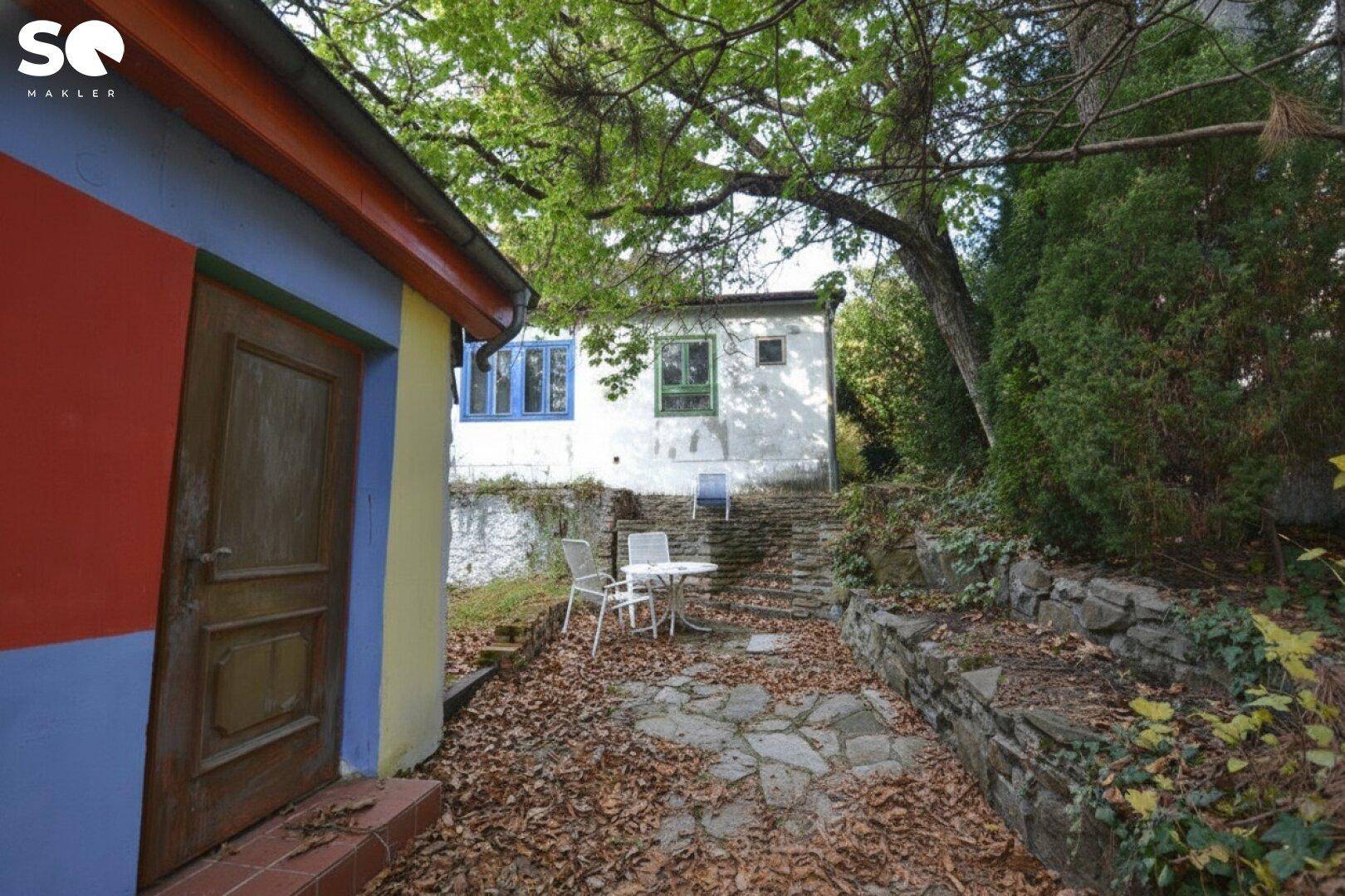 Rustikaler Außenbereich mit Steinfliesen, Gartenmöbeln und einem bunten Nebengebäude.