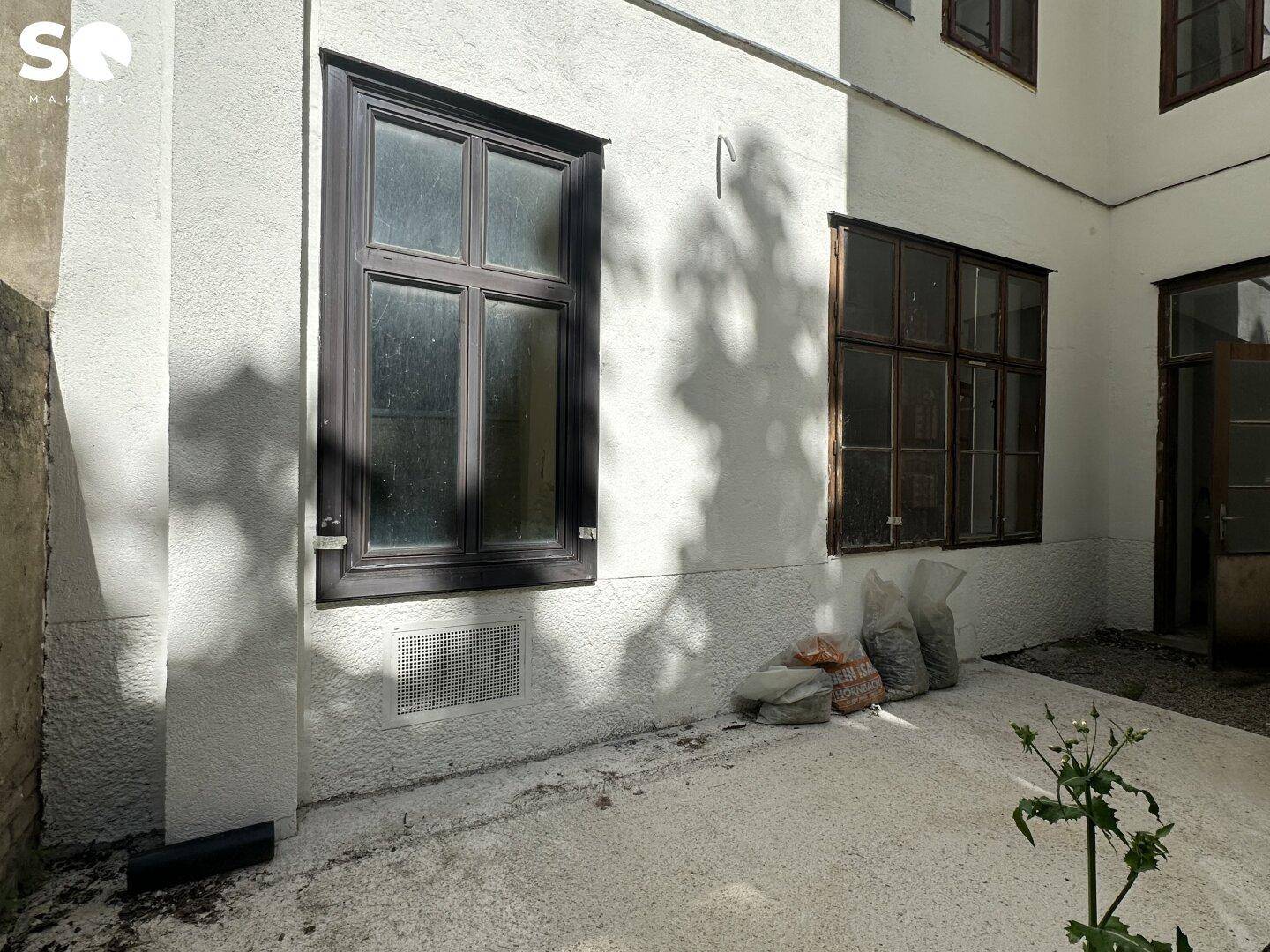 Heller Innenhof mit gepflastertem Boden, dunklen Fenstern und einem kleinen Pflanzenbeet.