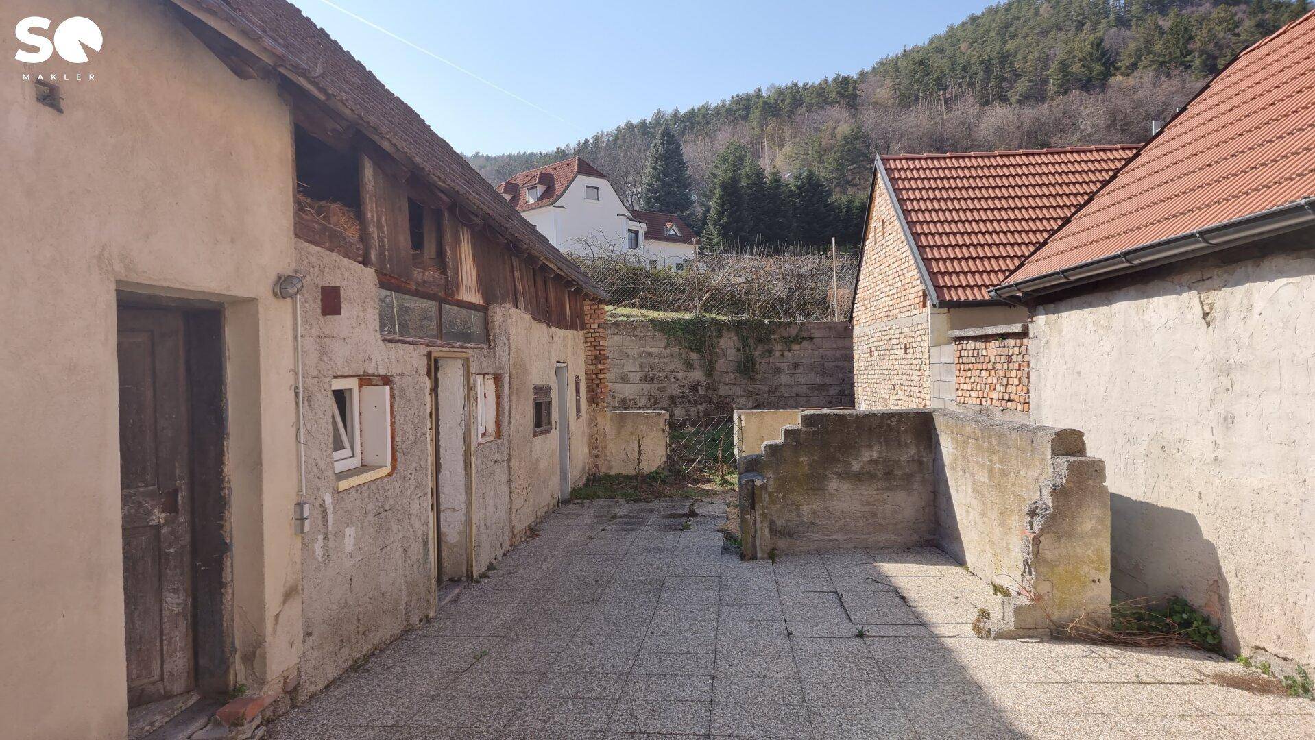 Alter Hofbereich mit Nebengebäuden und Blick auf bewaldete Hügel.