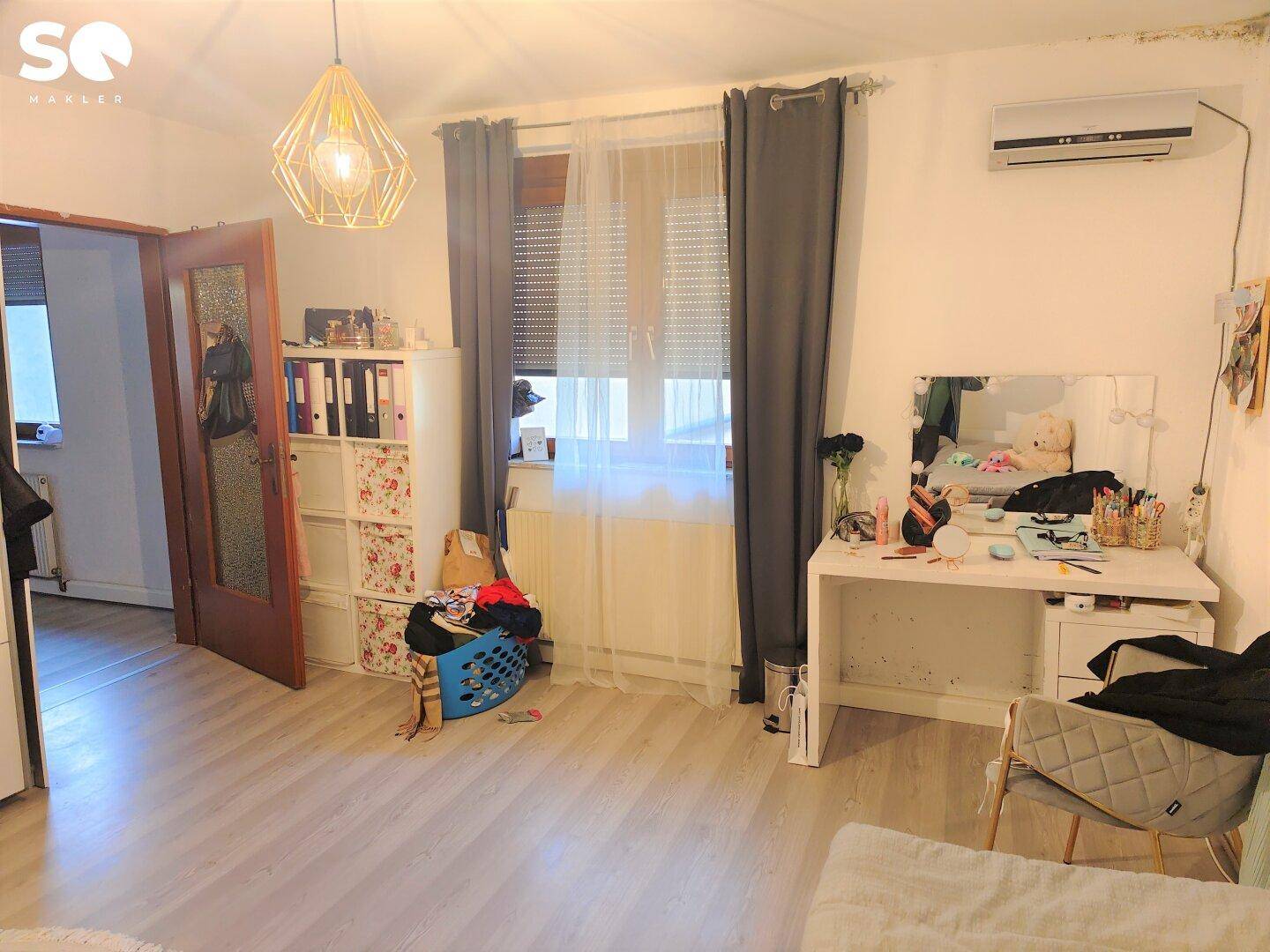 Schlafzimmer mit Fenster, Schminktisch und Klimaanlage an der Wand.