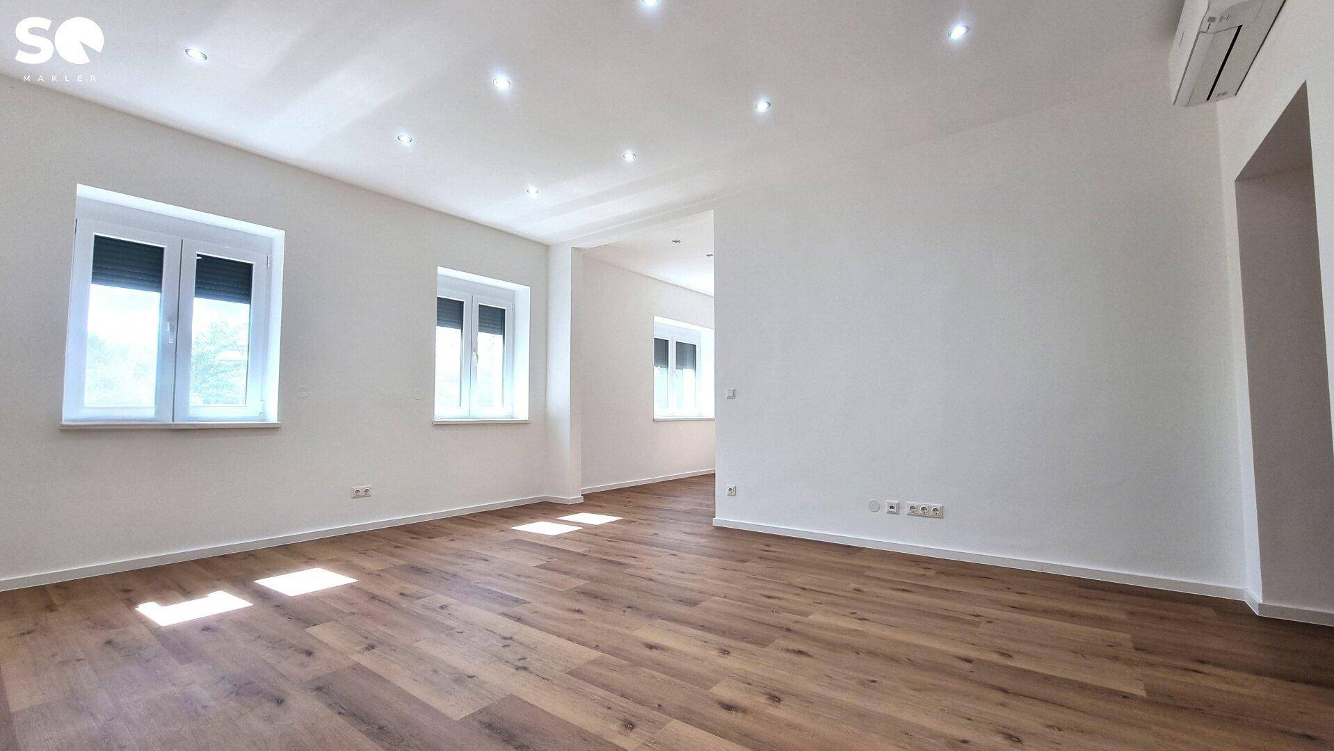 Lichtdurchfluteter Wohnbereich mit großen Fenstern, Laminatboden und Einbaustrahlern.