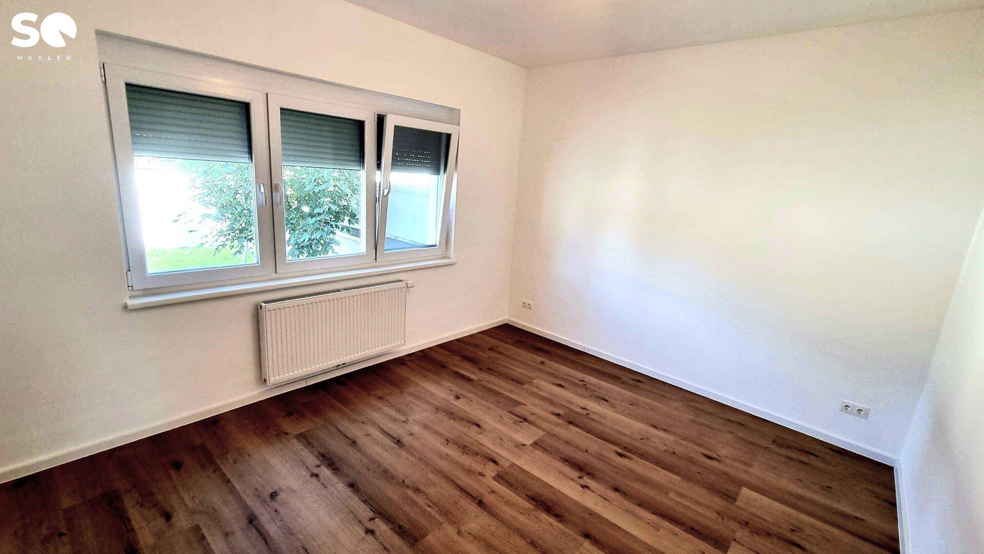 Helles Schlafzimmer mit Laminatboden, großem Fenster und Heizkörper unter dem Fenster.