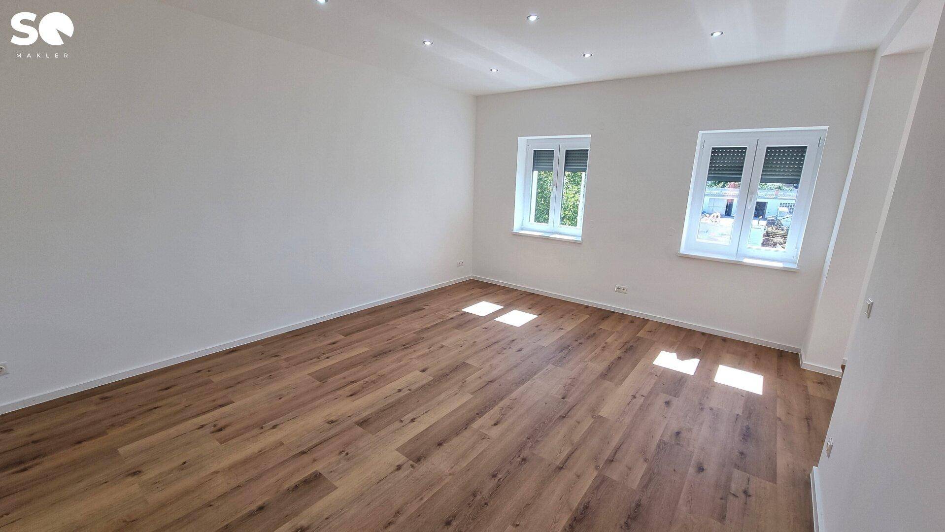 Heller, leerer Wohnraum mit Laminatboden, Einbaustrahlern und zwei Fenstern.