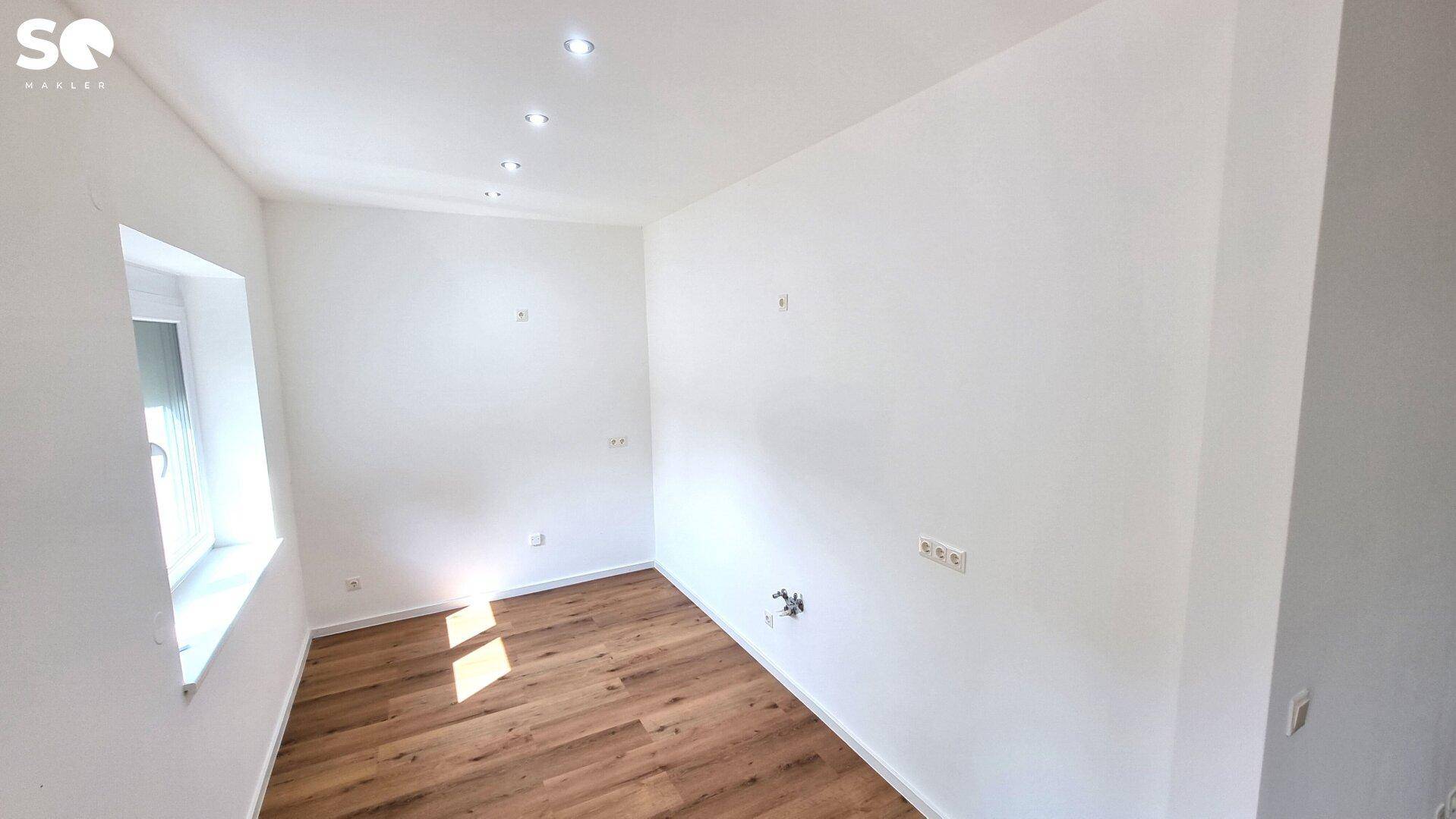 Heller Raum mit Laminatboden, Fenster und sichtbaren Anschlüssen an der Wand.