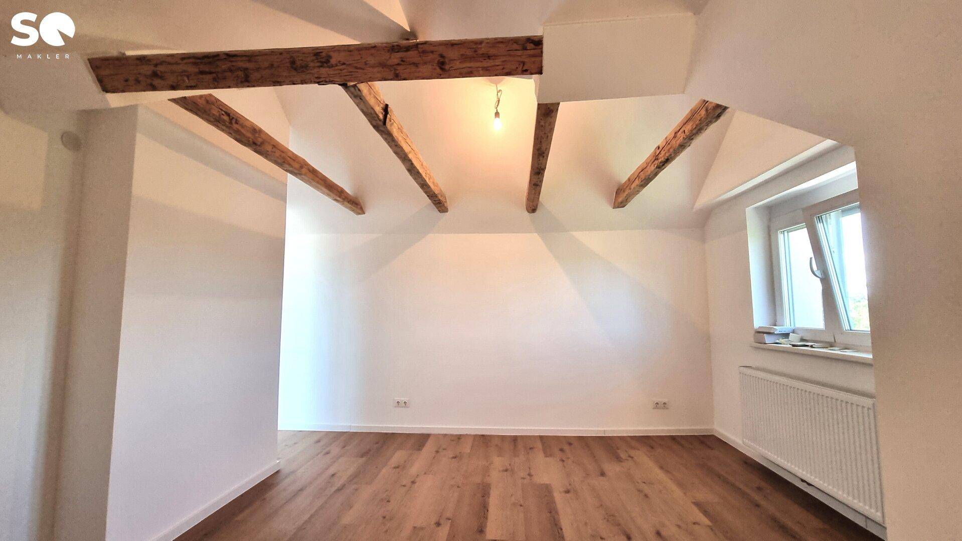 Geräumiger Dachbodenraum mit sichtbaren Holzbalken, Holzboden und einem Fenster für natürliches Licht.