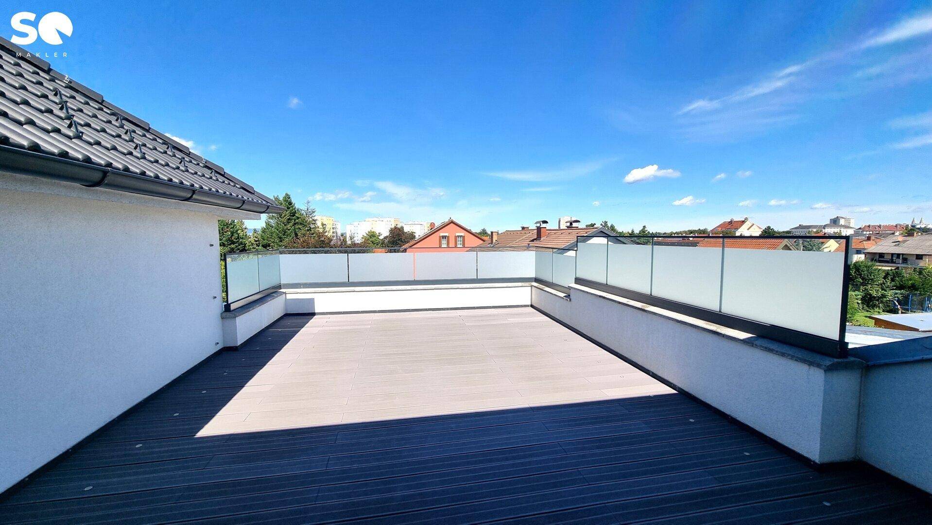 Geräumige Dachterrasse mit Holzdielenboden und Glasgeländer unter strahlend blauem Himmel.