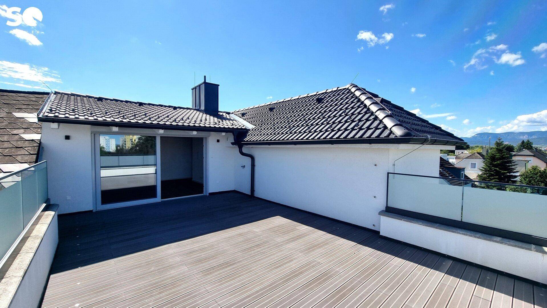 Weitläufige Dachterrasse mit Holzdielen und Glasgeländer, angrenzend an das Haus mit Ziegeldach.