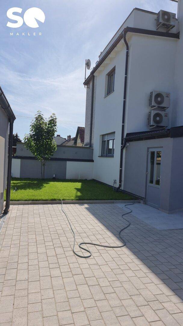 Seitenansicht eines modernen Hauses mit gepflasterter Zufahrt, Rasenfläche und Klimaanlagen.