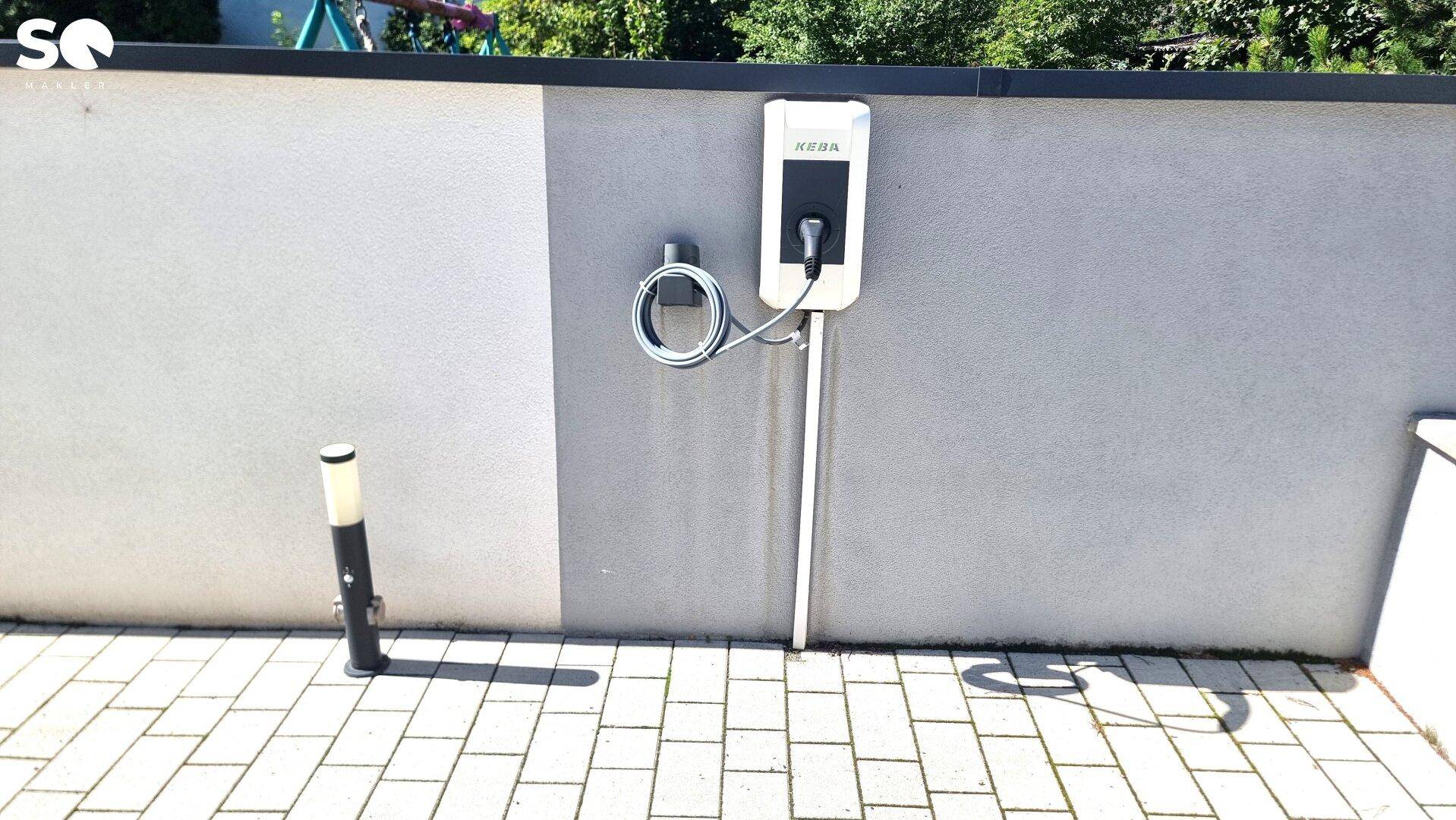 Ladestation für Elektrofahrzeuge an einer Wand neben einem gepflasterten Bereich.