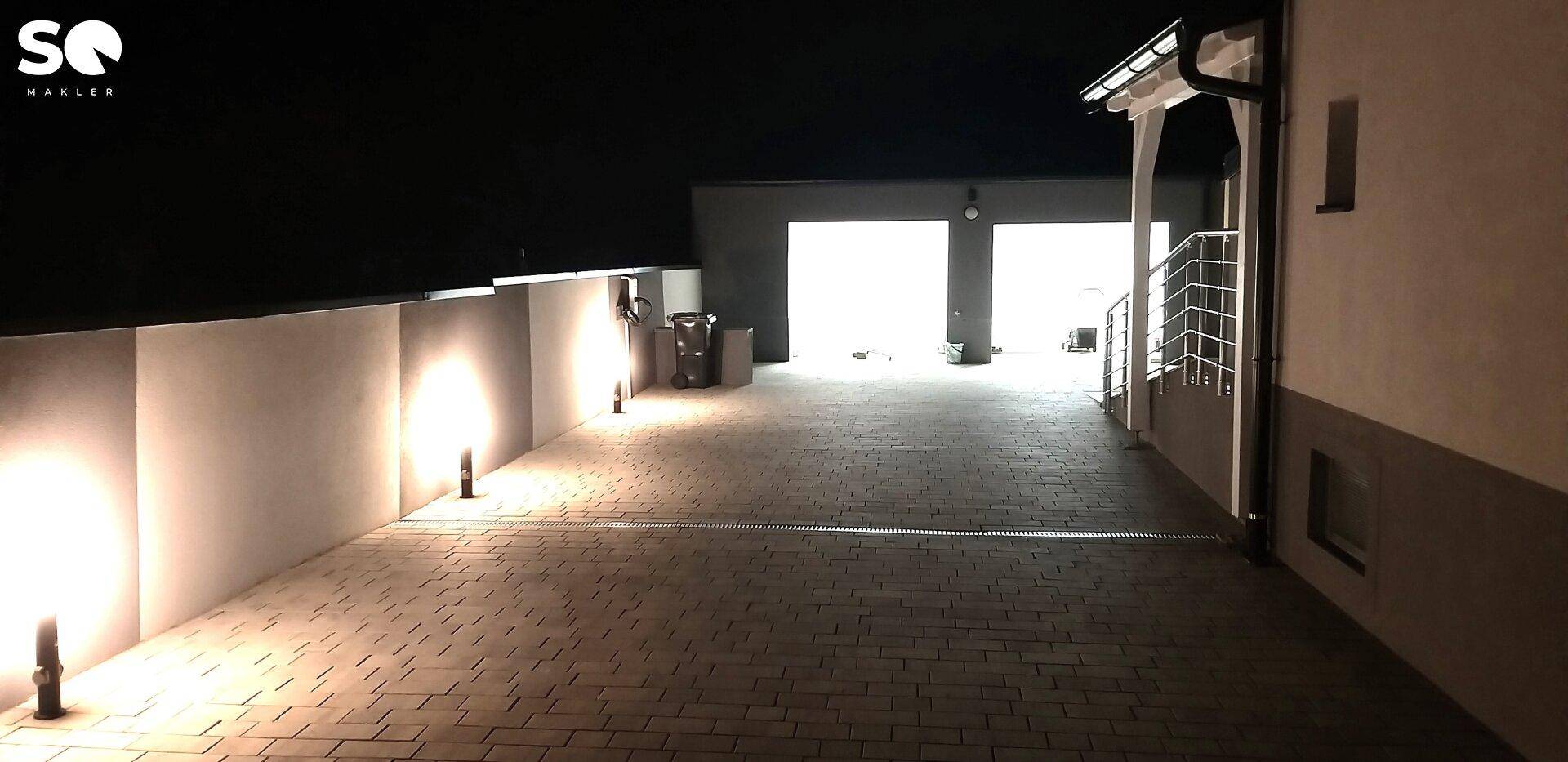 Beleuchtete Zufahrt und Garage bei Nacht, akzentuiert durch Bodenleuchten.