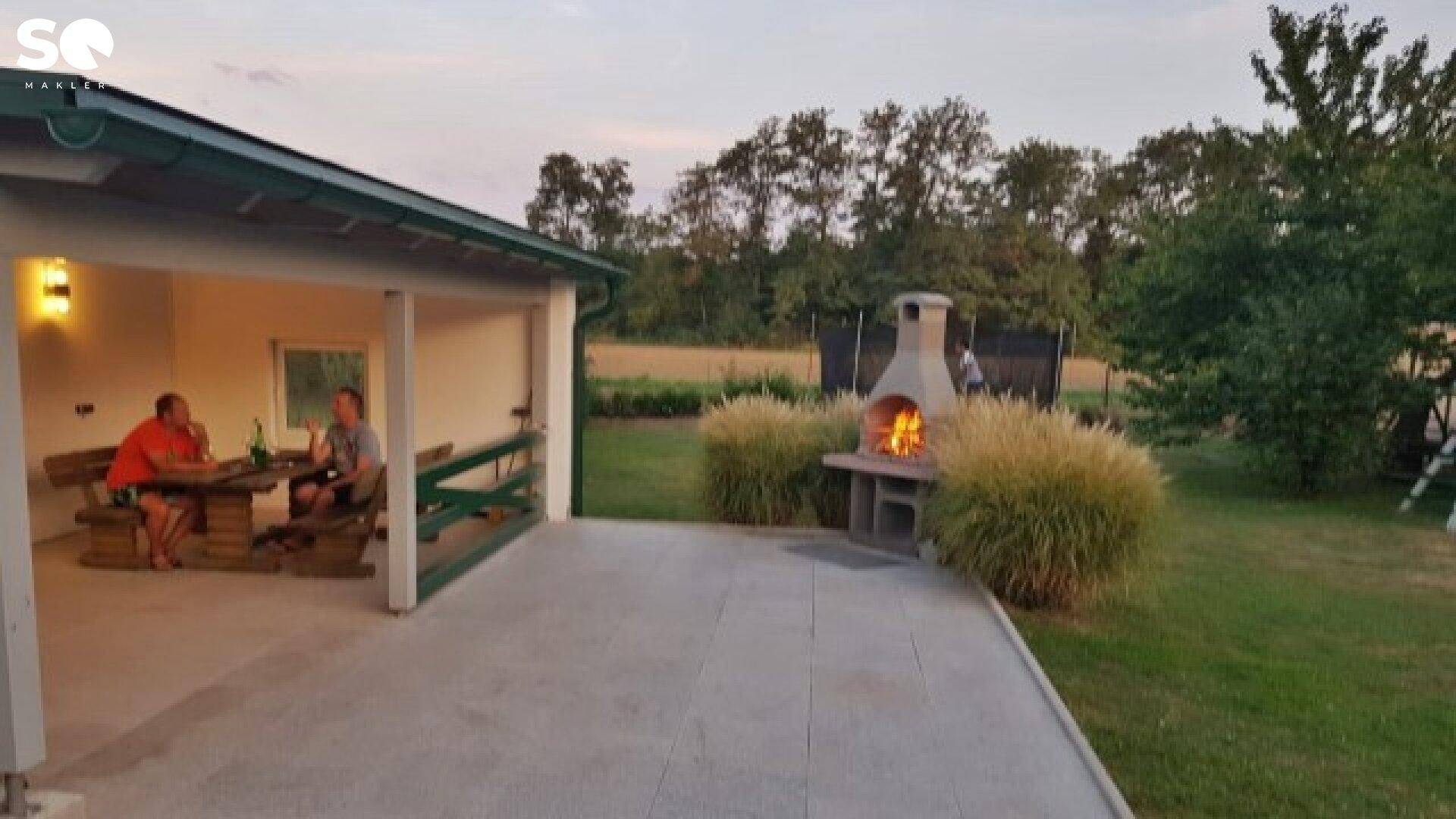 Überdachte Terrasse mit Sitzgelegenheiten und gemauertem Grillplatz im Garten.