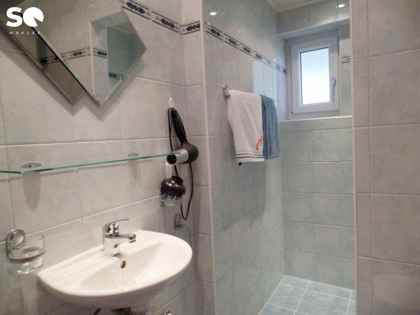 Gefliestes Badezimmer mit Dusche, Waschbecken und Fenster.