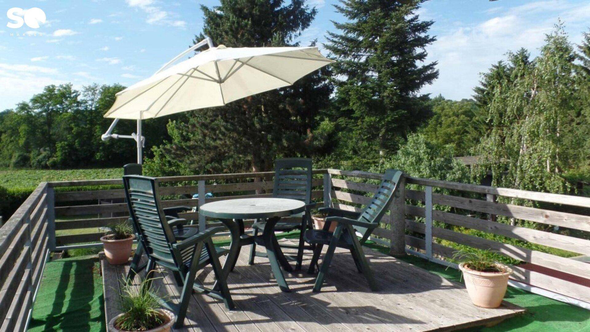 Holzterrasse mit Gartenmöbeln und Sonnenschirm, umgeben von Bäumen.