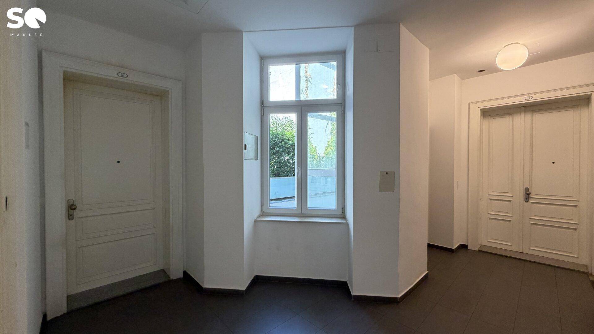 Heller Flur mit dunklem Fliesenboden und einem Fenster mit Blick ins Grüne.