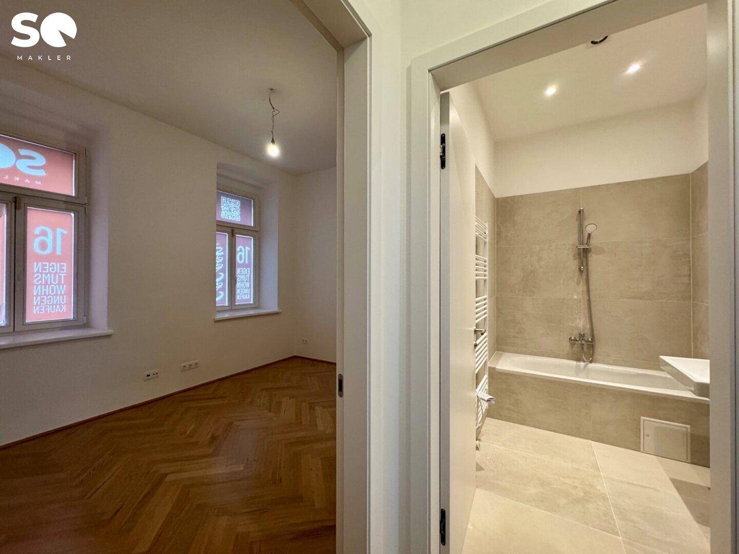 Blick aus einem Zimmer mit Fischgrätparkett in das angrenzende, modern geflieste Badezimmer.