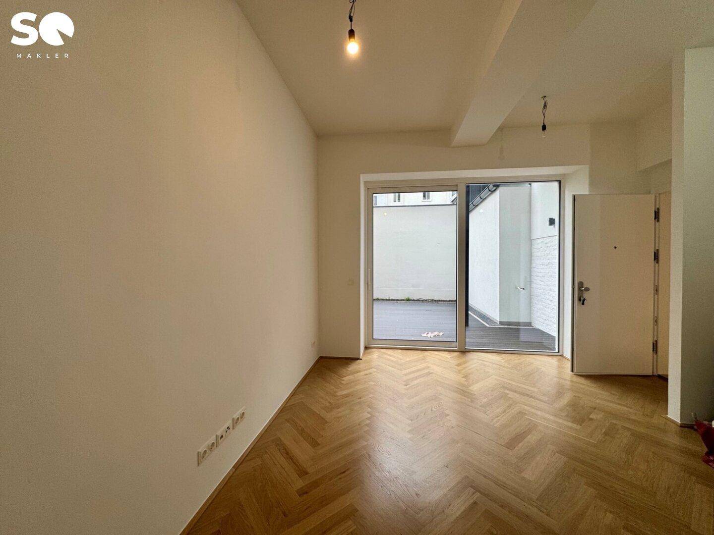 Geräumiger Wohnbereich mit Fischgrätparkett, Eingangstür und großem Schiebefenster zur Terrasse.