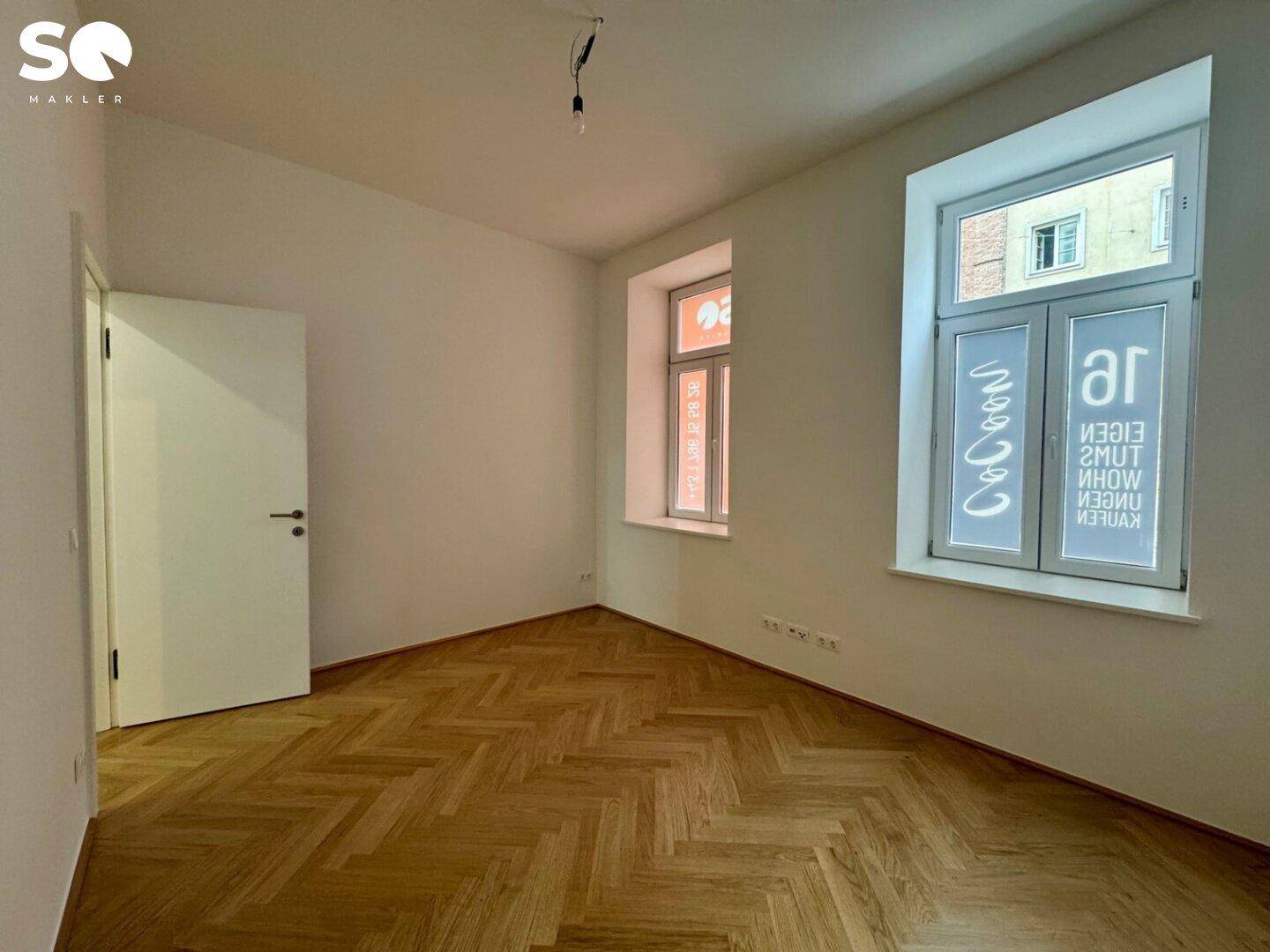 Geräumiges Zimmer mit klassischem Fischgrätparkett und zwei Fenstern, die viel Tageslicht hereinlassen.