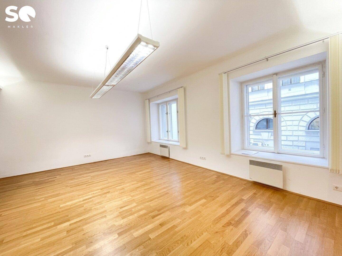 Heller, leerer Raum mit Holzparkettboden und zwei großen Fenstern, die viel Tageslicht hereinlassen.