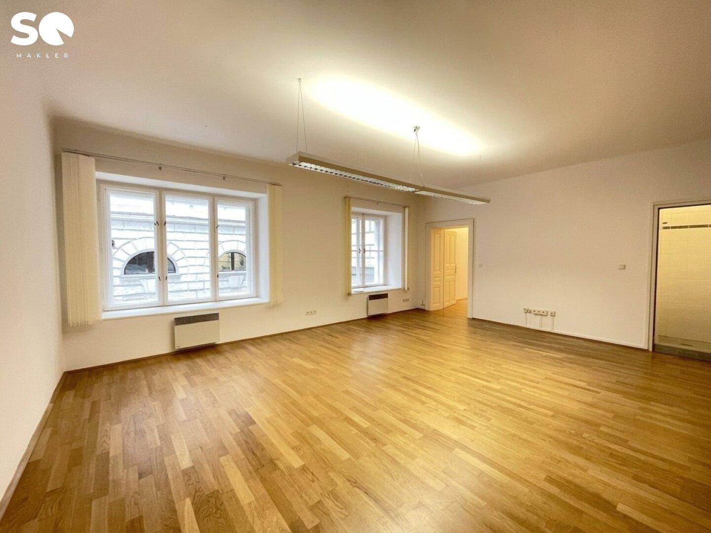 Geräumiger, heller Raum mit Holzboden und Fenstern, die den Blick auf die gegenüberliegende Architektur freigeben.