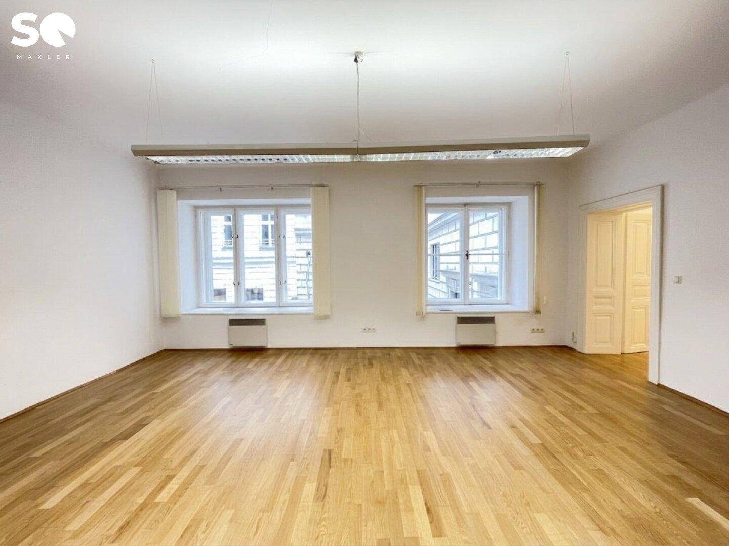 Großer, heller Raum mit Parkettboden und zwei Fenstern, die den Blick auf die umliegenden Gebäude ermöglichen.