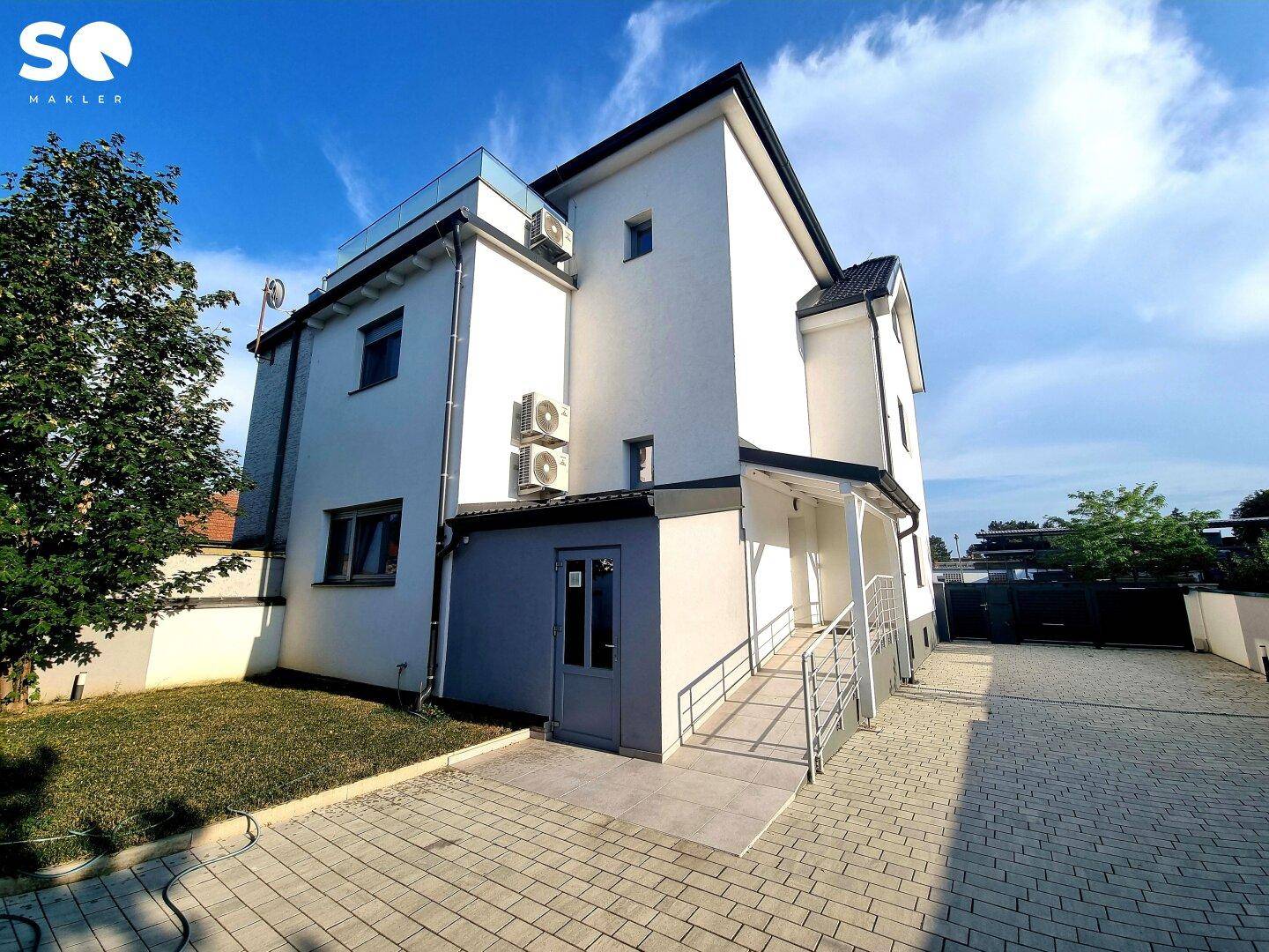 Modernes weißes Gebäude mit gepflasterter Auffahrt, kleiner Rasenfläche und barrierefreiem Zugang über eine Rampe.