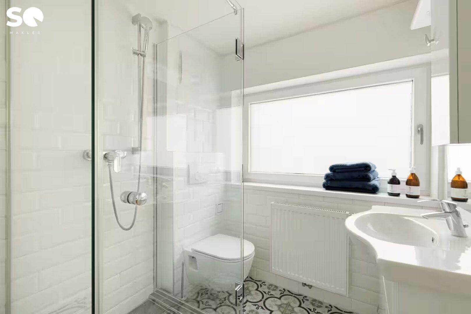 Helles Badezimmer mit Duschkabine, Fenster, modernem Waschbecken und gemusterten Bodenfliesen.