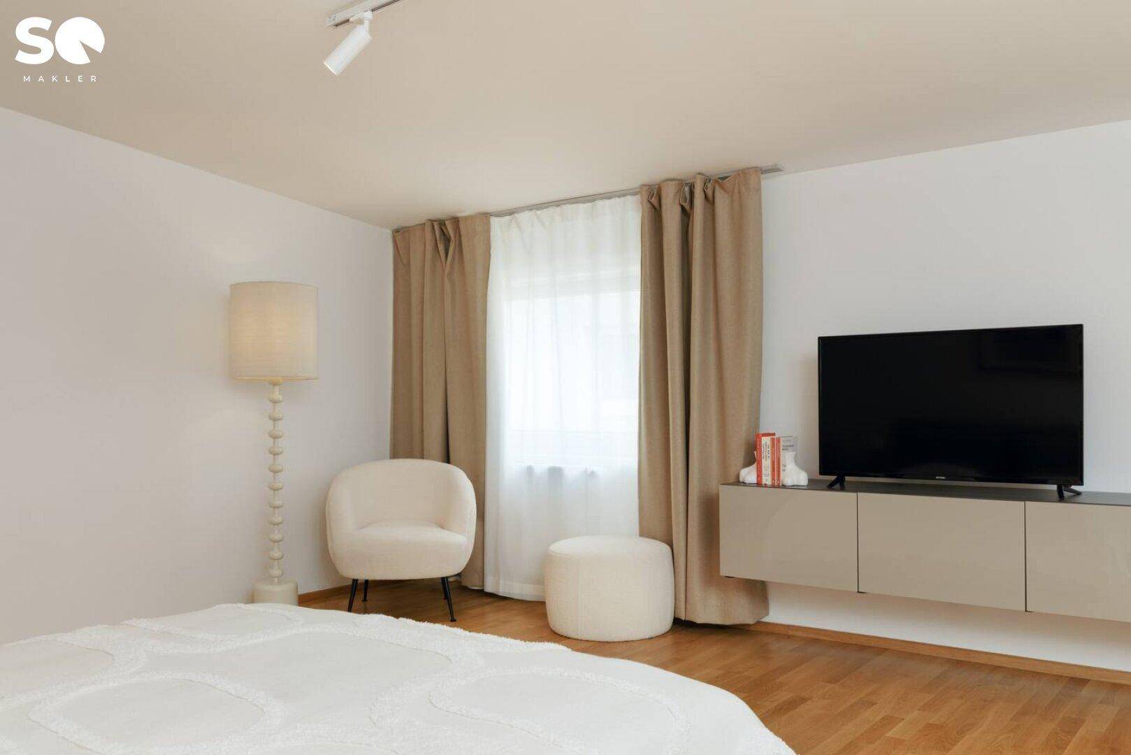 Helles Schlafzimmer mit Fernseher, bequemen Sitzgelegenheiten und Fenster mit Vorhängen.