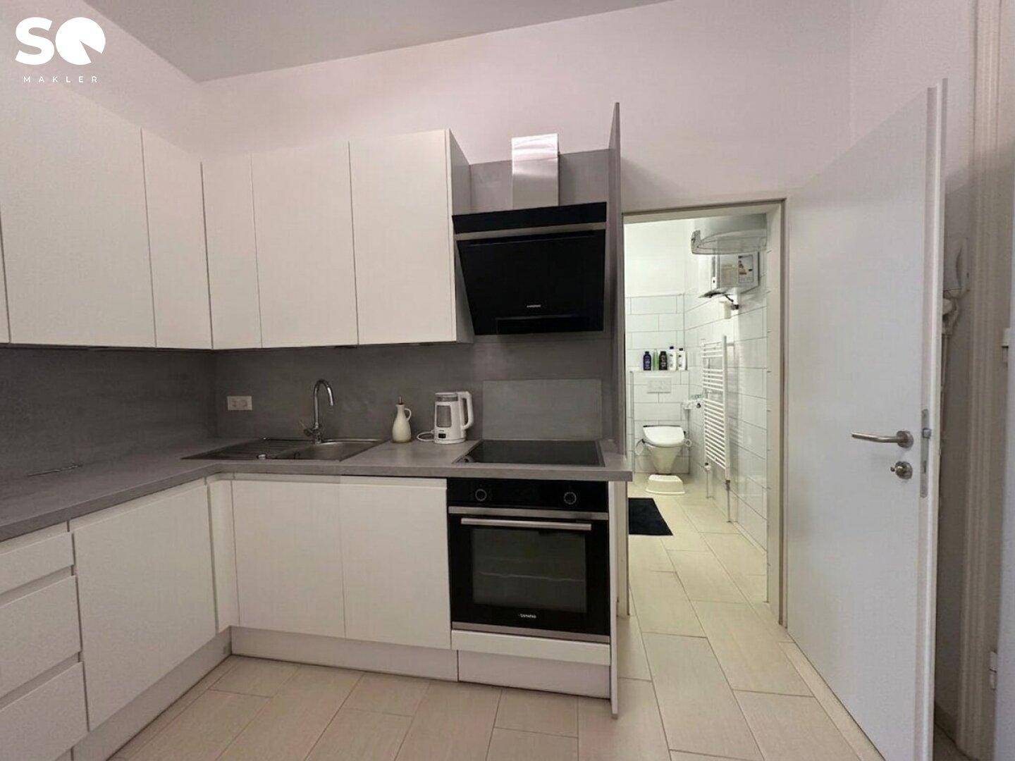Moderne Einbauküche mit weißen Schränken, grauer Arbeitsplatte und direktem Zugang zum Badezimmer.