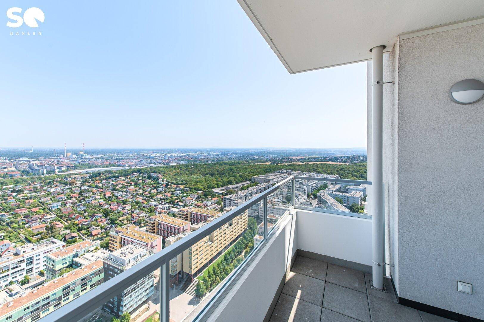 Großer Balkon mit Glasgeländer bietet einen weiten Panoramablick über die Stadt und grüne Flächen.