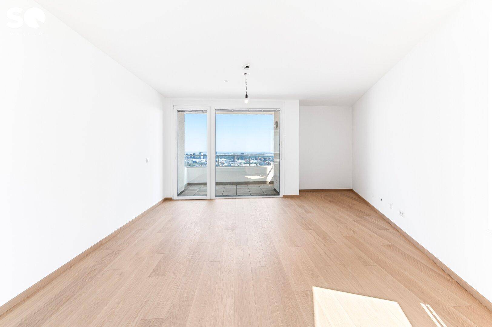 Geräumiges, helles Zimmer mit Holzboden und breiter Glasfront, die zu einem Balkon führt.