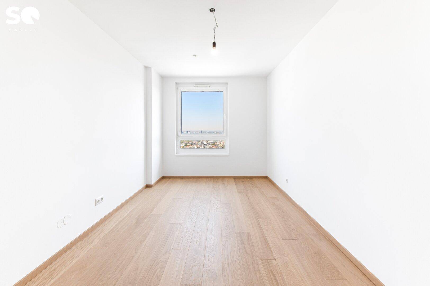 Helles, leeres Zimmer mit Holzboden und einem Fenster, das einen weiten Blick über die Stadt bietet.