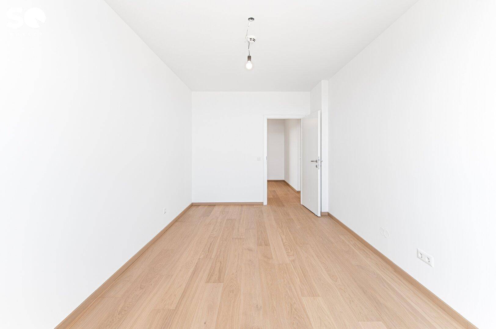 Leerer, heller Raum mit Holzboden und einer offenen Tür, die zu einem weiteren Bereich führt.