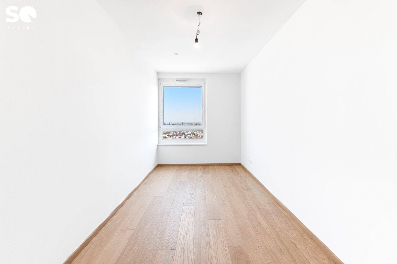 Leeres, helles Zimmer mit Holzboden und einem Fenster, das einen Panoramablick auf die Stadt ermöglicht.