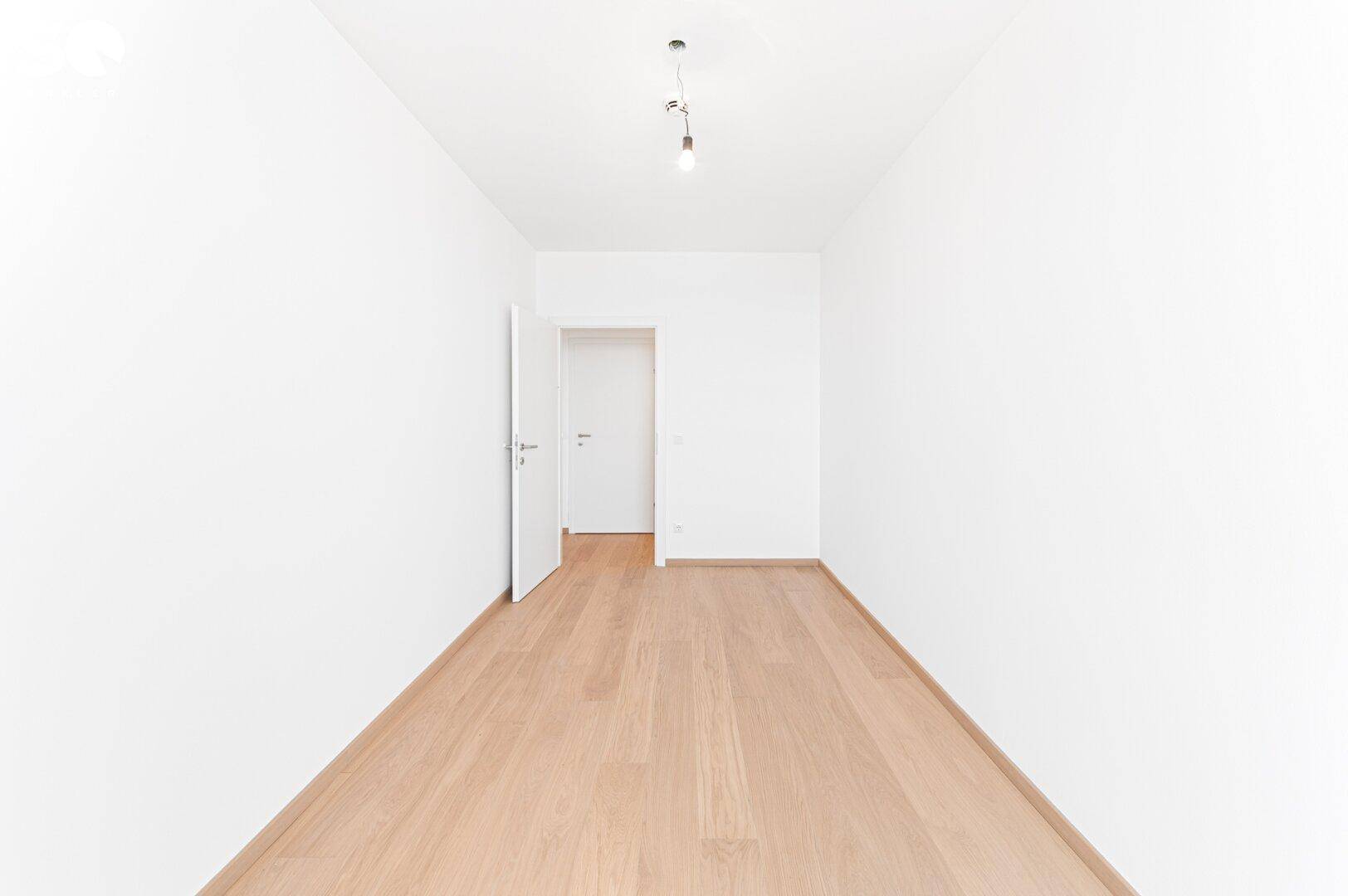 Heller, leerer Raum mit Holzboden und einer offenen Tür, die den Zugang zu anderen Zimmern zeigt.