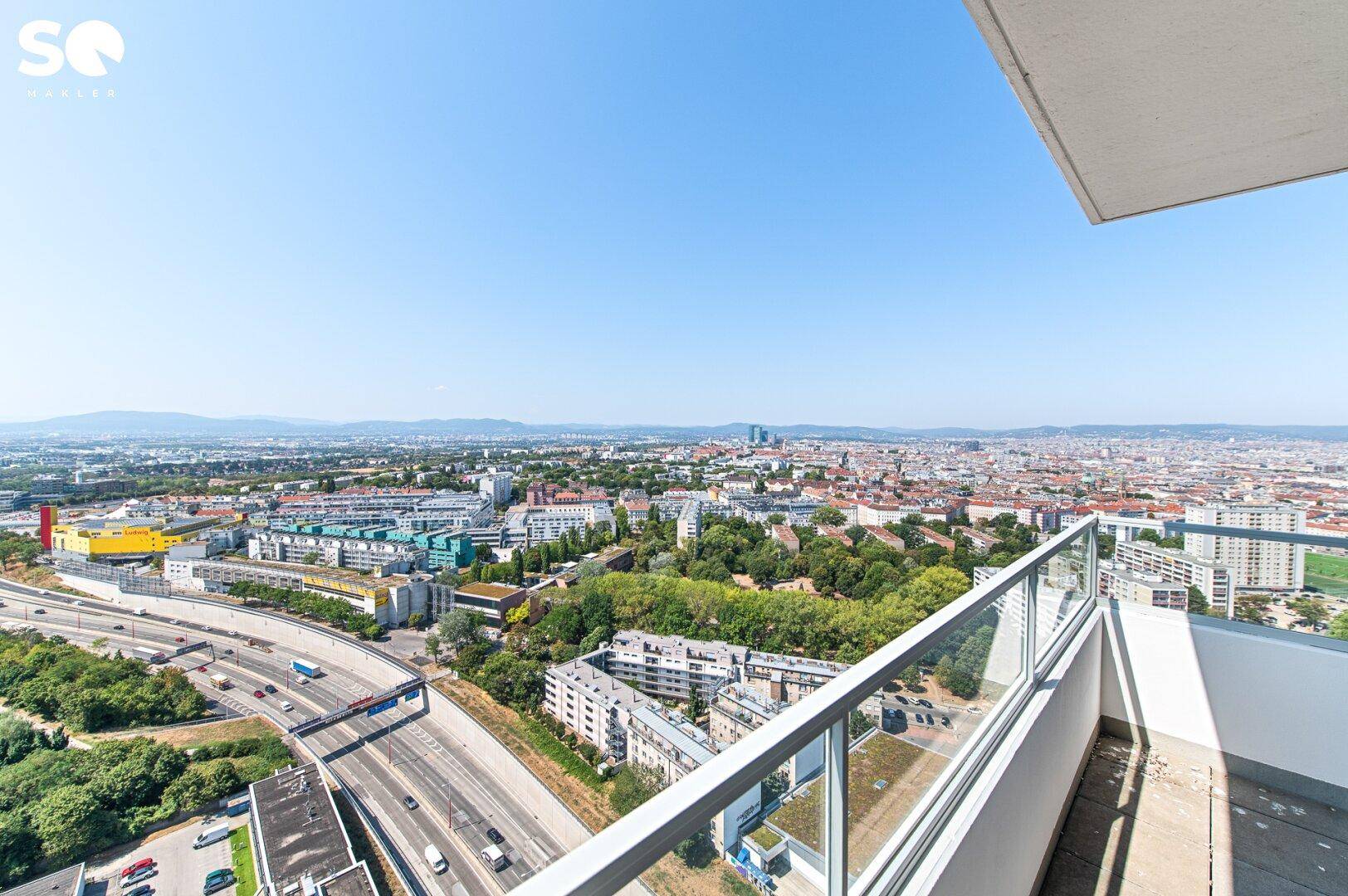 Weitläufiger Stadtblick von einem Balkon mit Autobahn, Gebäuden und Bergen am Horizont.