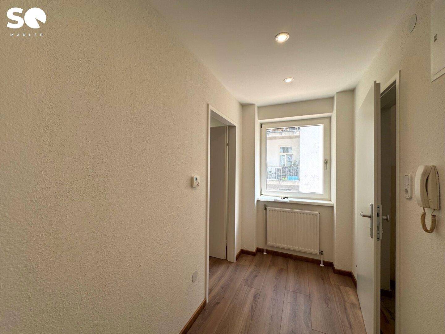 Heller Flur mit Laminatboden, Fenster und Gegensprechanlage, der zu weiteren Räumen führt.