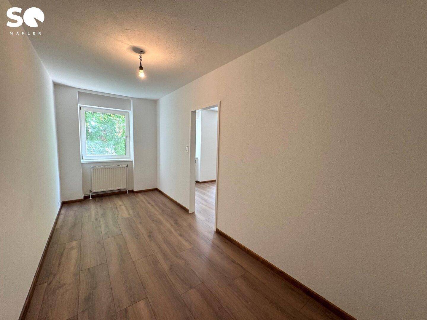 Helles Zimmer mit Laminatboden und einem Fenster, das einen Blick ins Grüne bietet.
