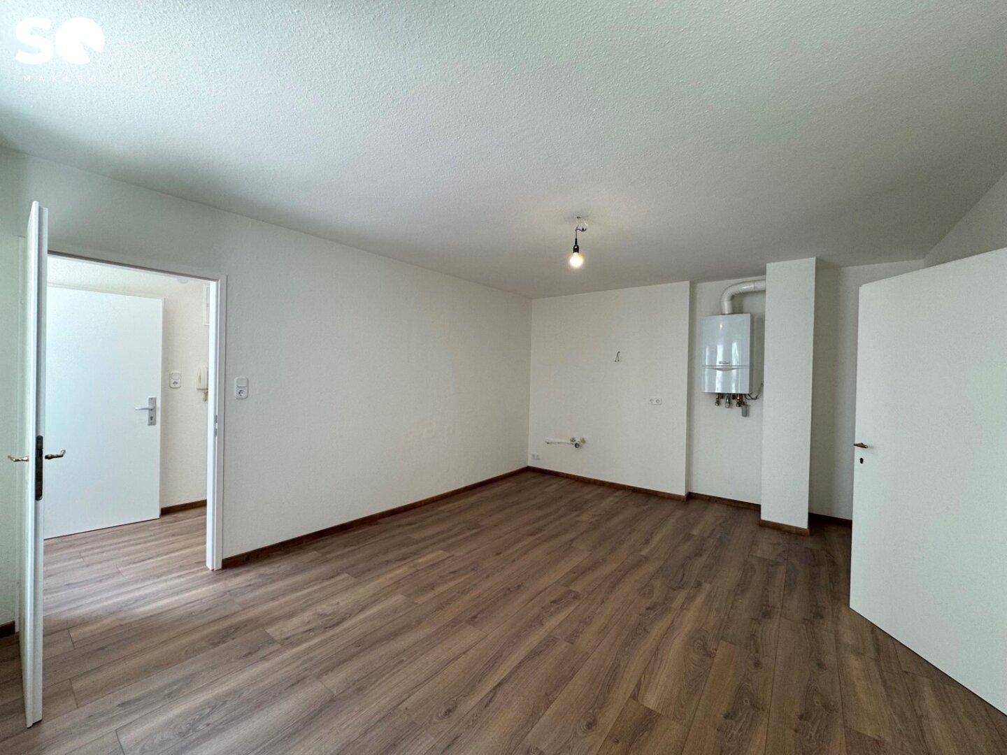 Leerer Raum mit Laminatboden und an der Wand montiertem Heizkessel, bereit für die Einrichtung.