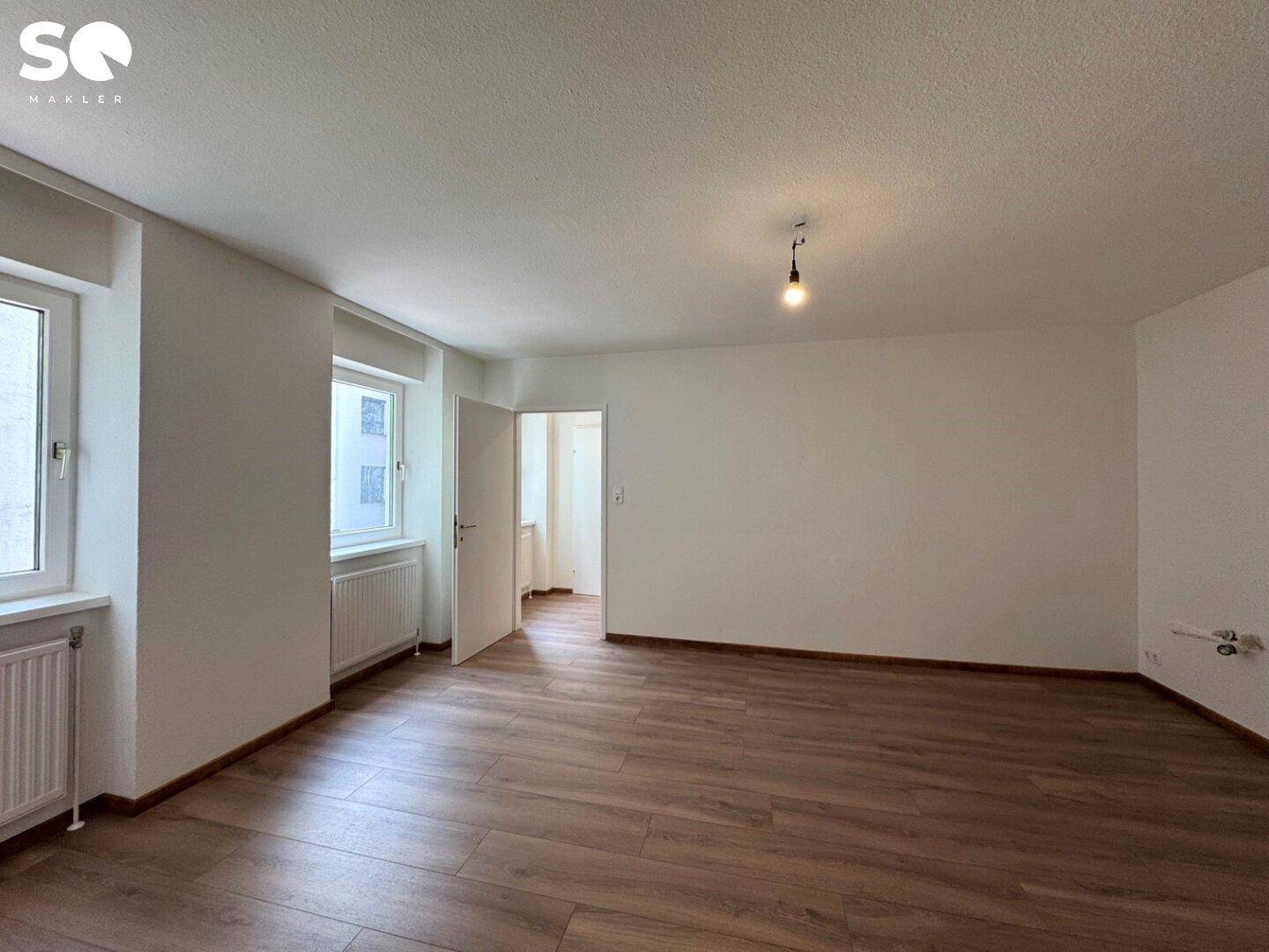 Großer, heller Raum mit Laminatboden und zwei Fenstern, die viel Tageslicht hereinlassen.