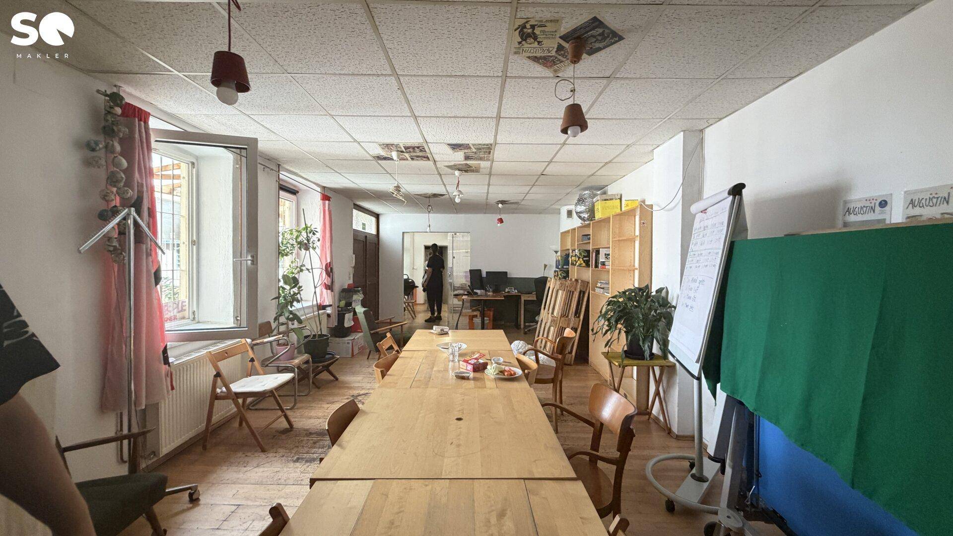 Langer Büroraum mit Holzboden, großen Fenstern und einem langen Tisch, der als Arbeitsbereich dient.