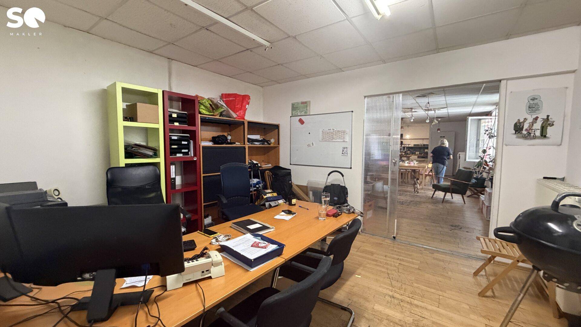 Büroarbeitsplatz mit Schreibtisch, Bürostühlen und Regalen, durch eine Glastür mit einem weiteren Raum verbunden.