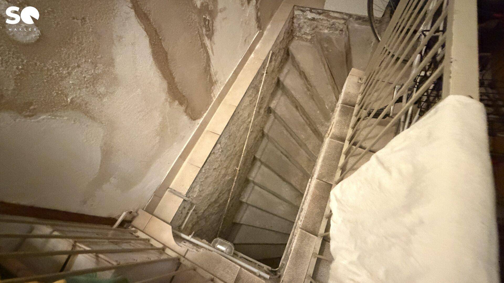 Enge Treppe mit gefliesten Stufen und einem Metallgeländer, die in ein Untergeschoss führt.
