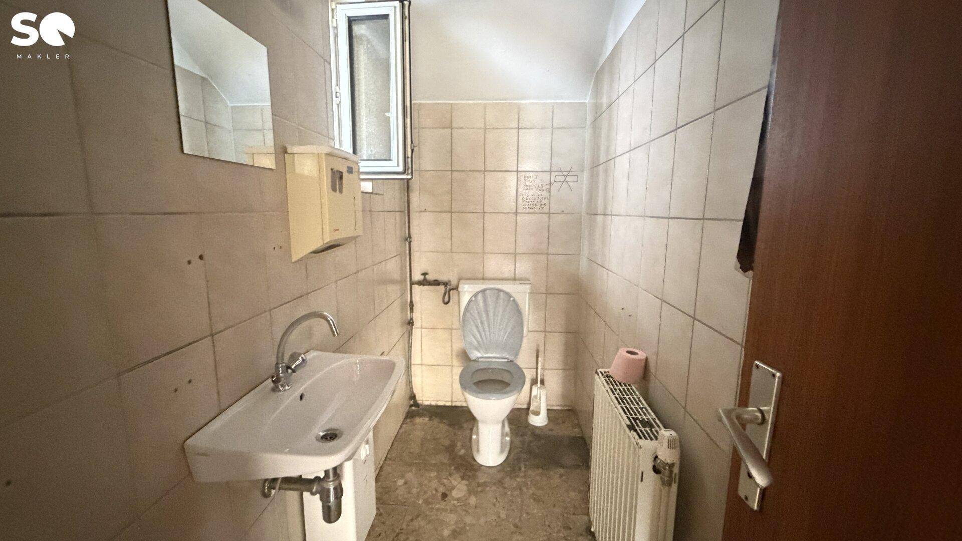 Kleine Toilette mit Waschbecken, Heizung und einem Fenster, gefliest in hellem Beige.
