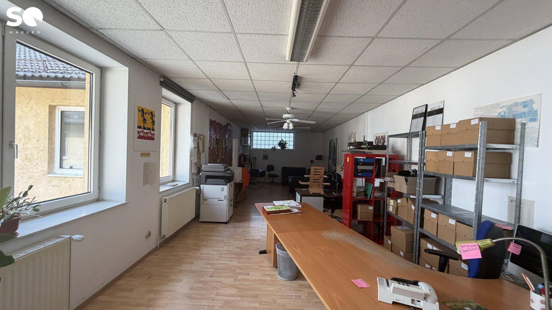 Langer Büroraum mit großen Fenstern, Holzboden und mehreren Arbeitsplätzen.