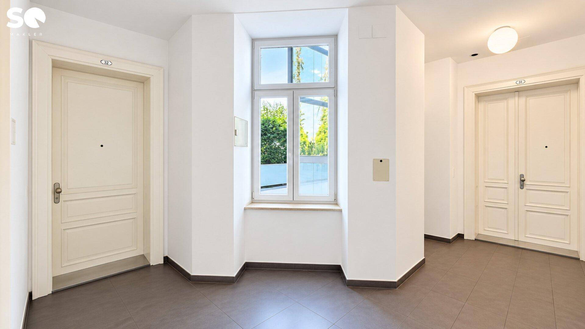 Heller Flur mit Fliesenboden, zwei Wohnungstüren und einem Fenster mit Blick ins Grüne.