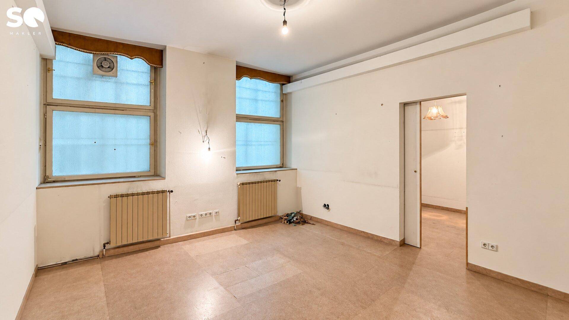Leerer Raum mit zwei Fenstern, Heizkörpern und einer einfachen Deckenleuchte.